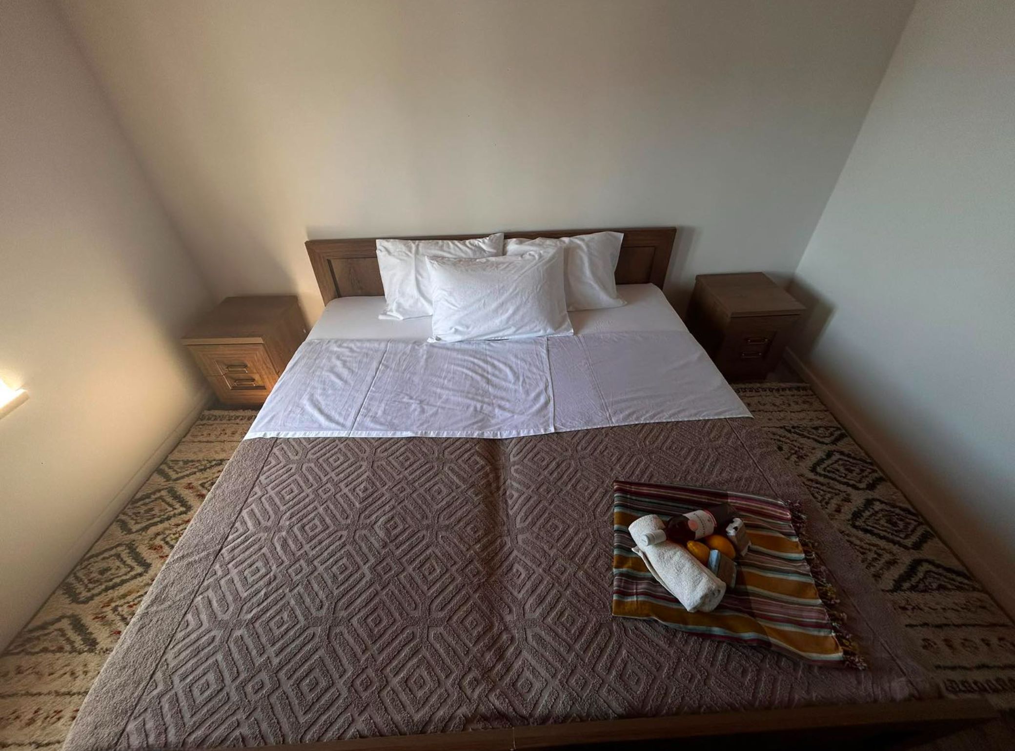 Een net opgemaakt bed met witte lakens en kussens, een kamerkleed met dessin, en een dienblad met handdoeken en toiletartikelen.