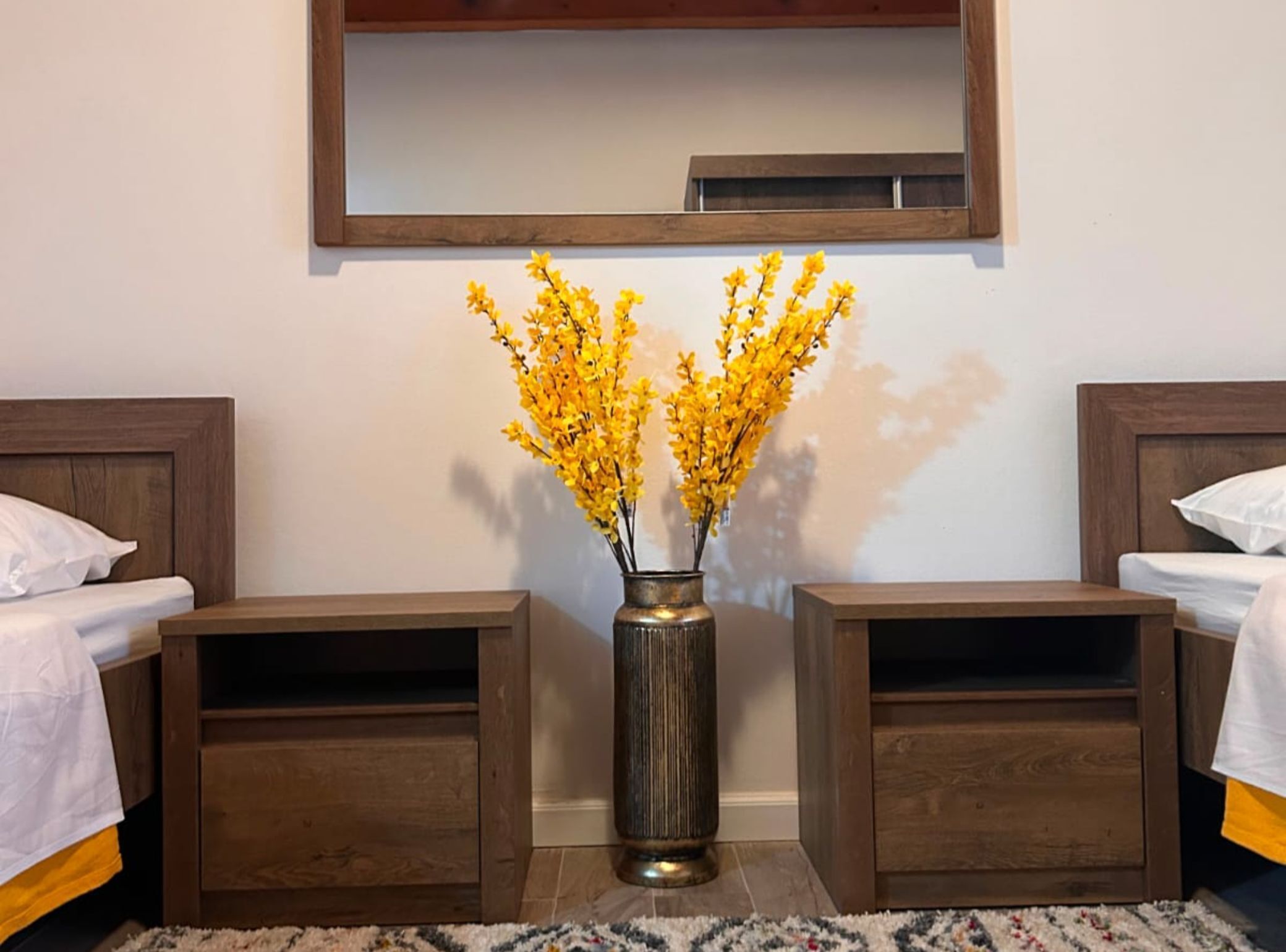 Quarto com duas mesas de cabeceira de madeira, um vaso central com flores amarelas e um espelho acima.