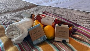 A bed with a striped blanket, a rolled white towel, a bottle of Alexandria wine, a box of Galebar soap, a box of Kenova soap, and two lemons.