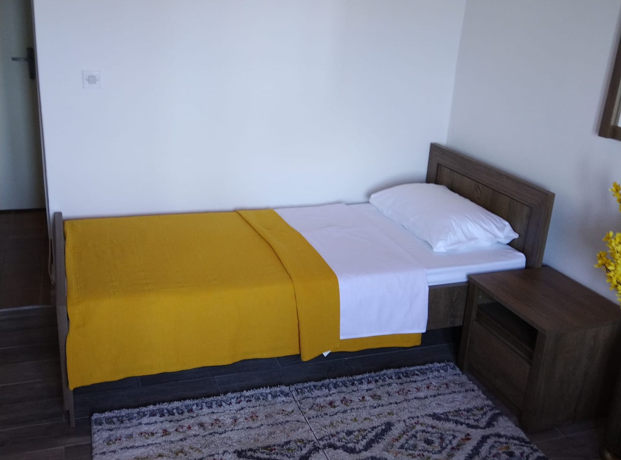 A single bed with a yellow bedspread and white linens, a wooden nightstand, a patterned rug, and a small wooden cabinet.