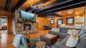 A cozy cabin interior with a stone fireplace, wooden floors, a gray sofa, and a dining area. - Epic Group Cabin | Game Room + King Suites + Views (Sevierville)