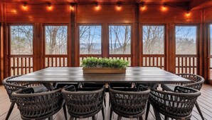 A wooden deck with a large table and wicker chairs, surrounded by windows with a view of trees. - Epic Group Cabin | Game Room + King Suites + Views (Sevierville)