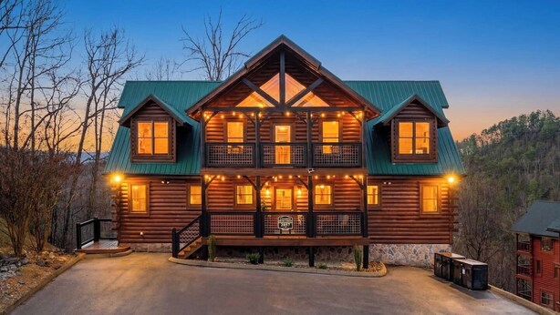 A multi-story log cabin with a balcony, a stone foundation, and a driveway leading to the entrance. - Epic Group Cabin | Game Room + King Suites + Views (Sevierville)