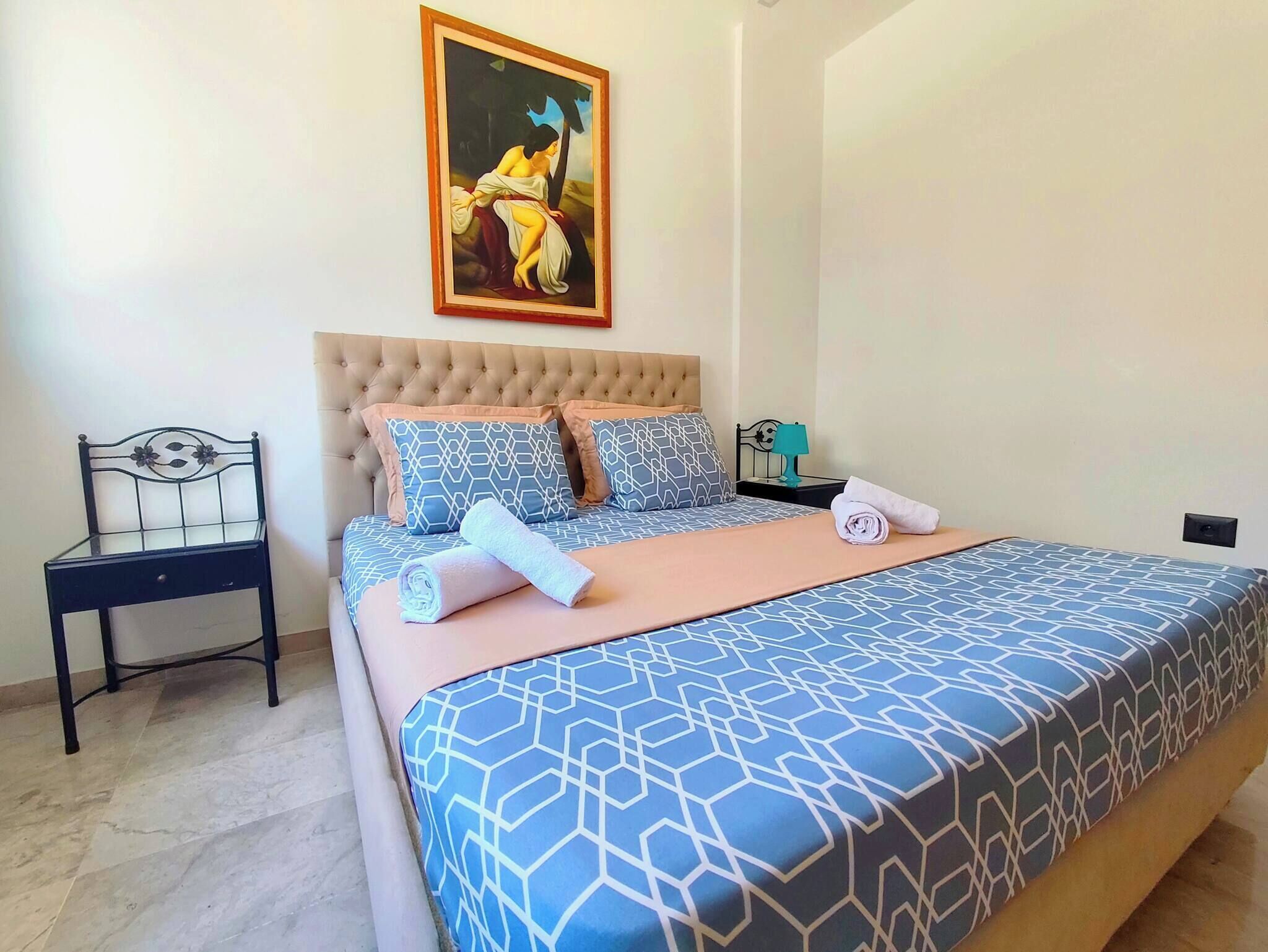 Een slaapkamer met een bed, een stoel, een nachtkastje, een lamp en een schilderij aan de muur.
