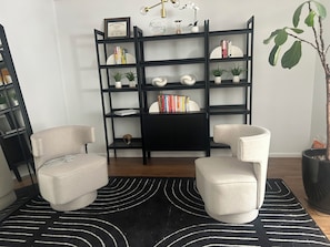 Ein Zimmer mit zwei Sesseln, einem schwarz-weißen Teppich, einem schwarzen Bücherregal mit Dekoration und einer Topfpflanze.