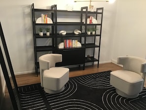 Een kamer met een zwarte schapunit, twee lichtgekleurde stoelen, een zwart-wit tapijt en een klein plantje op de planken.
