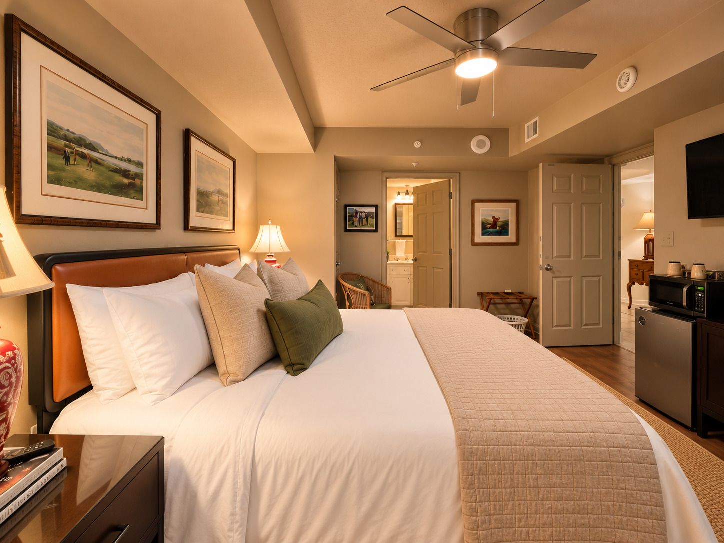 A hotel room with a bed, bedside table, lamp, ceiling fan, artwork on the walls, and a kitchenette area.