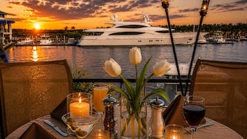 Une table dressée pour un repas, avec des bougies, un vase rempli de fleurs et des verres de vin, donnant sur une marina où sont amarrés des bateaux, le tout baigné par les couleurs d’un coucher de soleil.