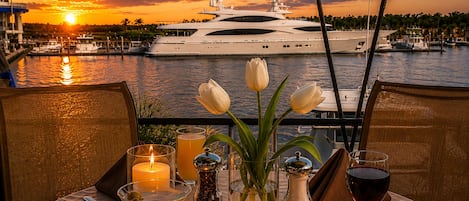 Mesa lista para una comida, con velas, un florero y copas de vino, con vista a un puerto deportivo con barcos y un atardecer.