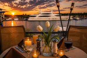 Une table dressée pour un repas, avec des bougies, un vase rempli de fleurs et des verres de vin, donnant sur une marina où sont amarrés des bateaux, le tout baigné par les couleurs d’un coucher de soleil.