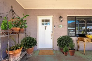 A front door with a doormat, potted plants, a metal shelf with more plants, a small table, and a window. - 3BR Near Hospitals & UofA w/ Workspace (Tucson)