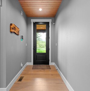 A hallway with a wooden floor, a black door, and a view of greenery outside. - Luxury Driggs Cabin • Grand Targhee • Jackson Hole • Tetons (Driggs)