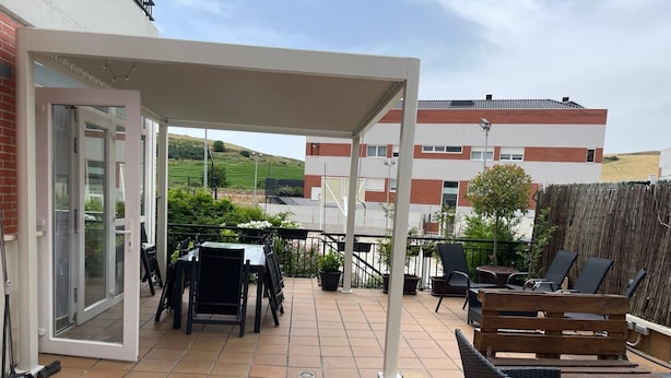 En overdækket terrasse med udendørs møbler, en skydedør og udsigt til et boligområde.