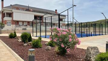 A residential building with a tennis court, surrounded by a fence, and a landscaped garden with rocks and shrubs.