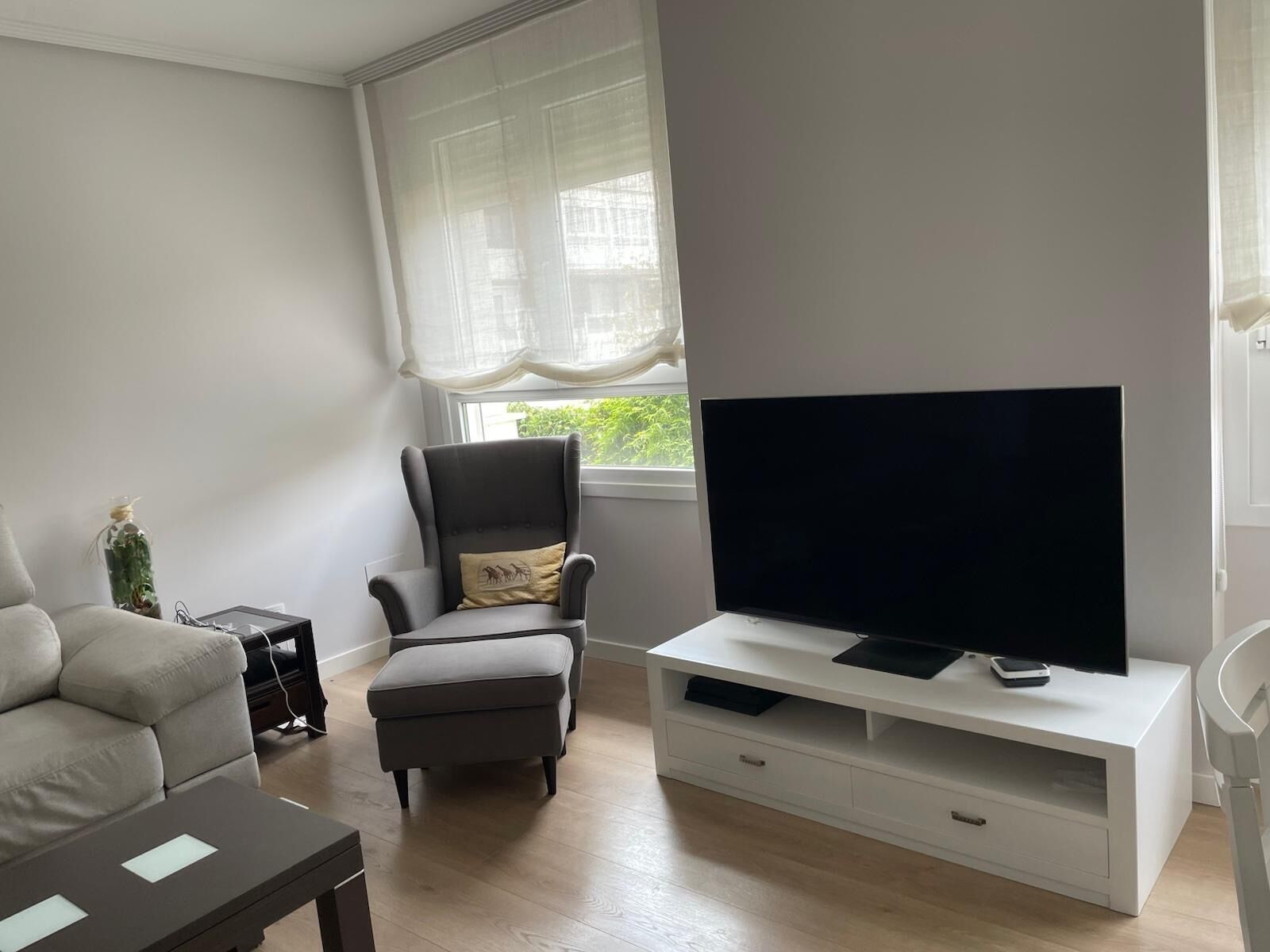 A living room with a sofa, a chair, a television on a white stand, and a window with sheer curtains.