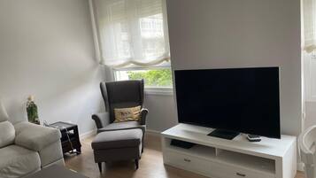 A living room with a sofa, a chair, a television on a white stand, and a window with sheer curtains.