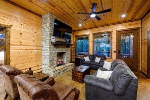 L’intérieur d’une cabane en rondins, avec une cheminée en pierre, des canapés en cuir, un ventilateur de plafond et de grandes fenêtres.