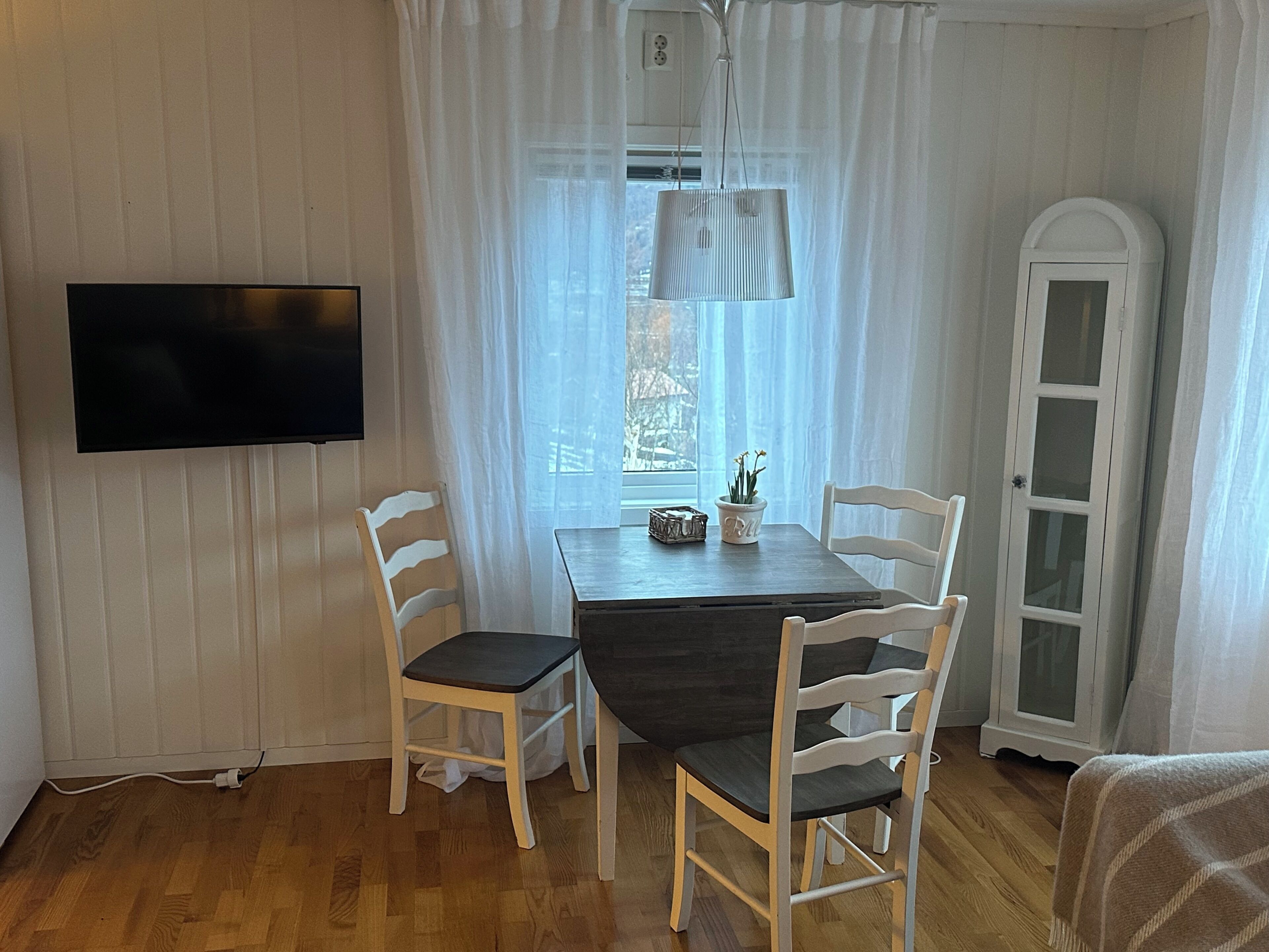 Un comedor con una mesa, sillas, un televisor, una ventana con cortinas y un armario.