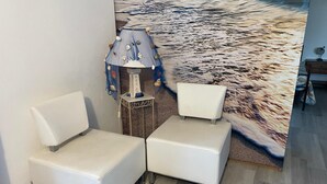 Habitació d'angle amb dues butaques de pell blanca, una taula petita amb una làmpada i un mural gran a la paret amb una escena de platja.