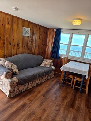 A room with a wooden paneled wall, a grey sofa with floral cushions, a small table with a white tablecloth, and a view of the sea through a window with blue curtains. - Wake Up to the Sound of Waves by the Shore in North Truro, Massachusetts (North Truro)