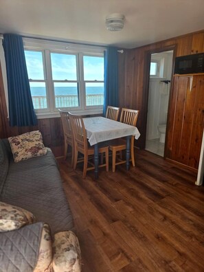 A room with a wooden floor, a dining table with chairs, a sofa with a floral pillow, and a view of the sea through large windows. - Wake Up to the Sound of Waves by the Shore in North Truro, Massachusetts (North Truro)