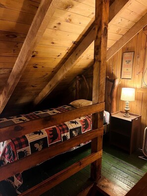 A rustic attic bedroom with a bunk bed, a wooden ceiling, and a nightstand with a lamp.