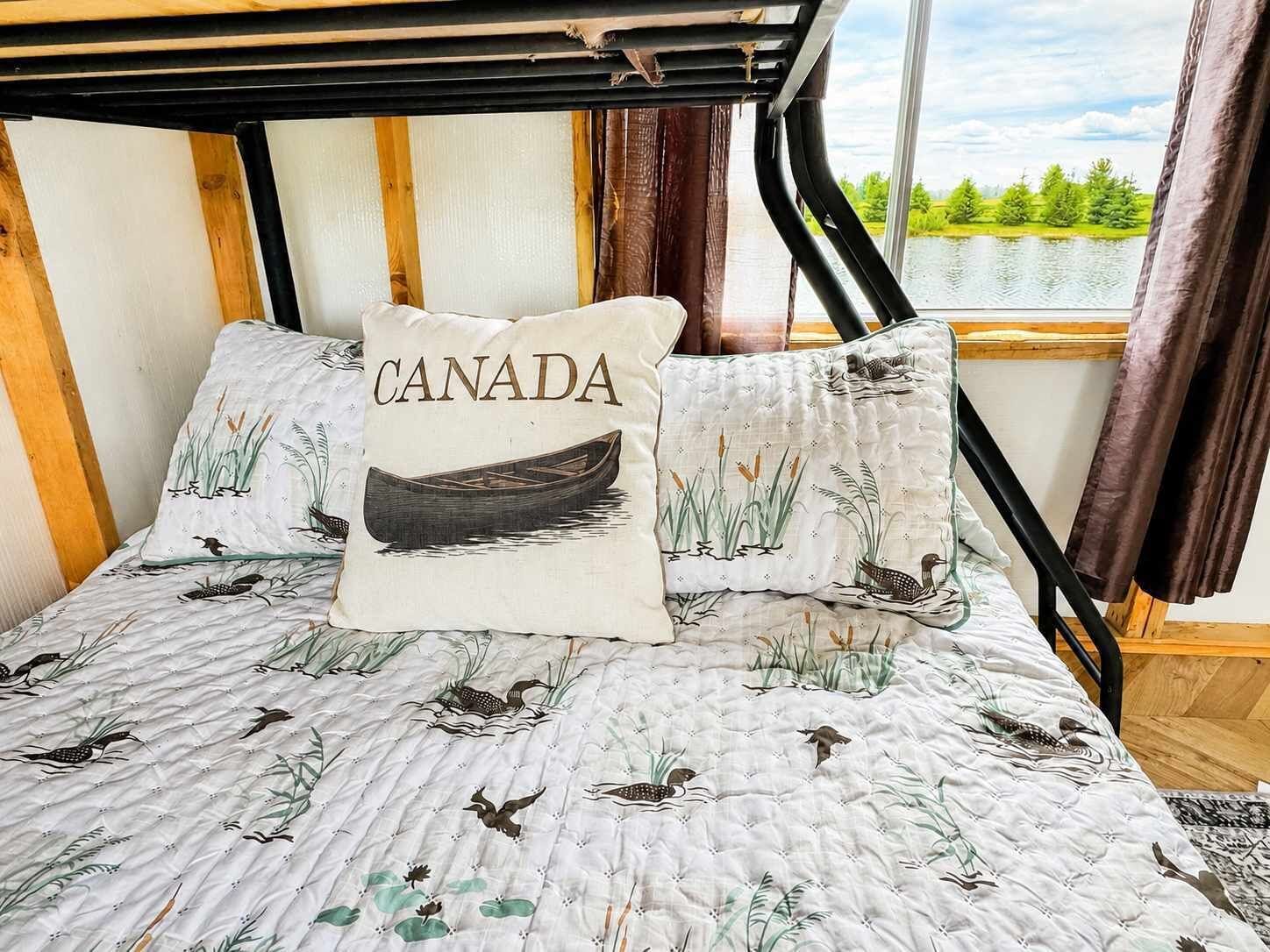 Ein Bett mit einem Kissen im kanadischen Design, Blick auf einen See und ein Fenster mit Vorhängen.