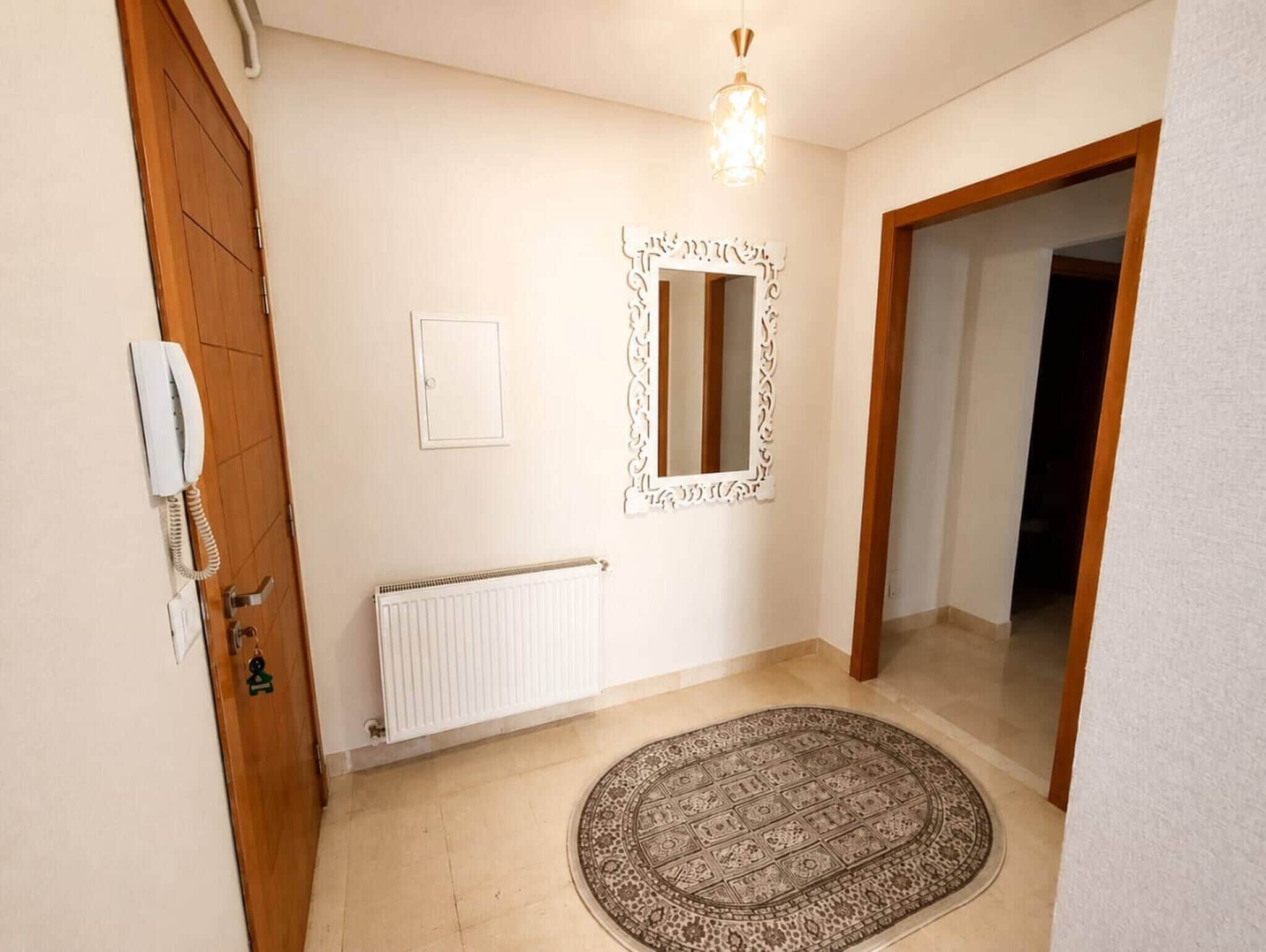 A hallway with a wooden door, a white radiator, a patterned rug, a mirror with an ornate frame, and a light fixture.