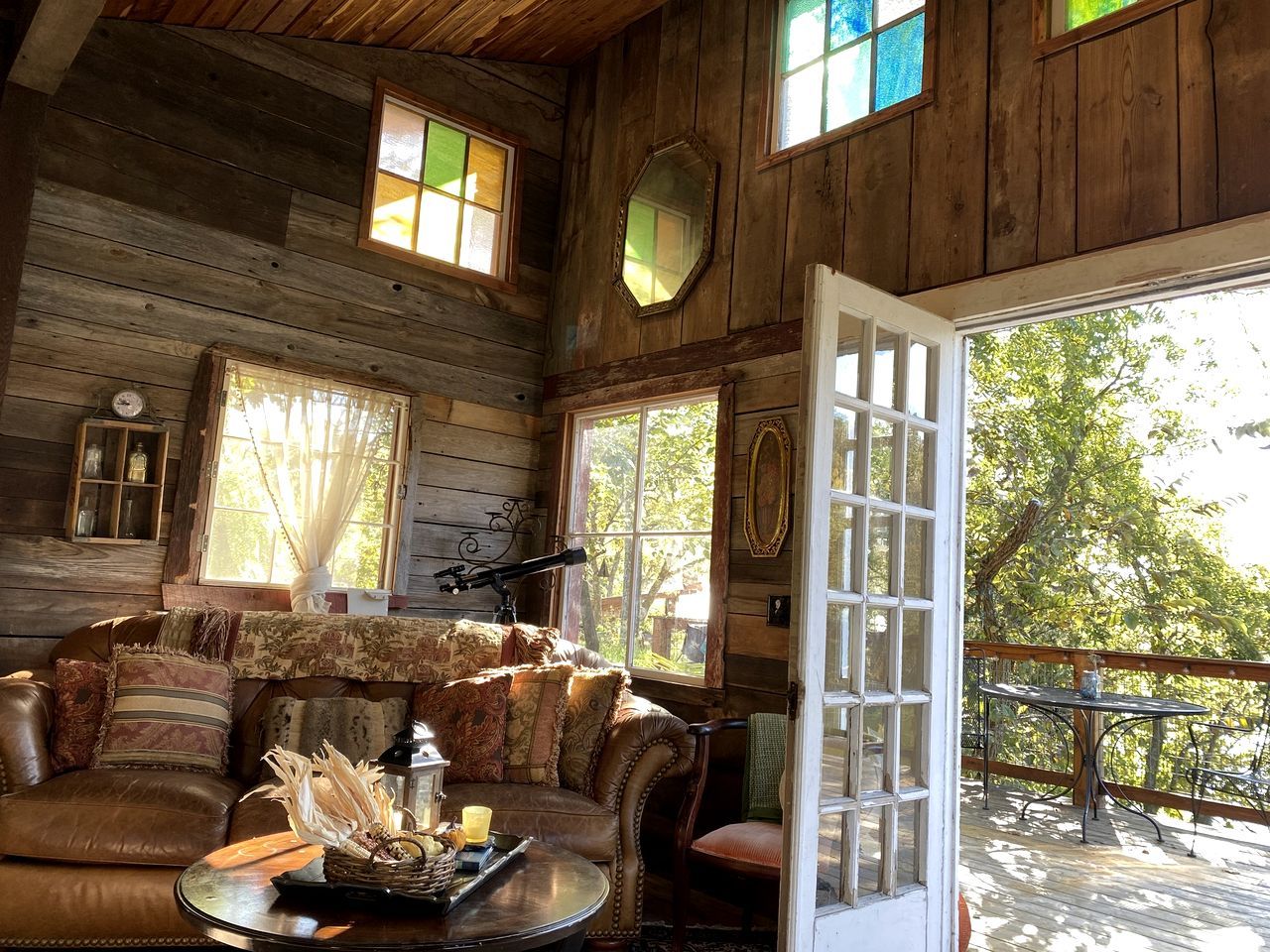 A rustic cabin interior with a leather sofa, a round table with a candle, and a large window with a view of trees.