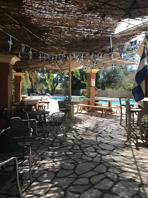 Área de piscina externa com piso de pedra, cadeiras de sol e área de estar coberta com bandeiras.