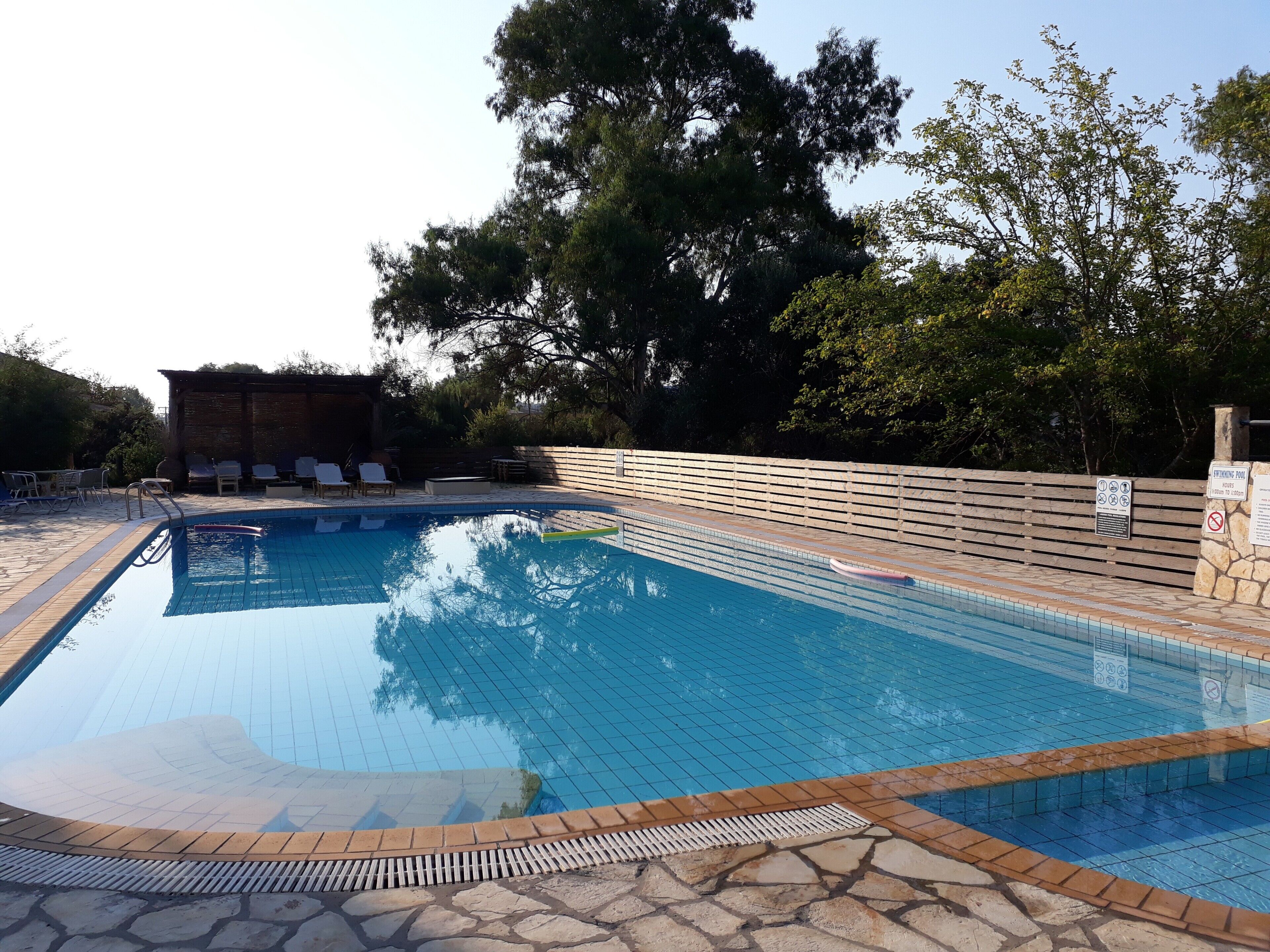 A swimming pool with a tiled deck, lounge chairs, and a wooden fence.
