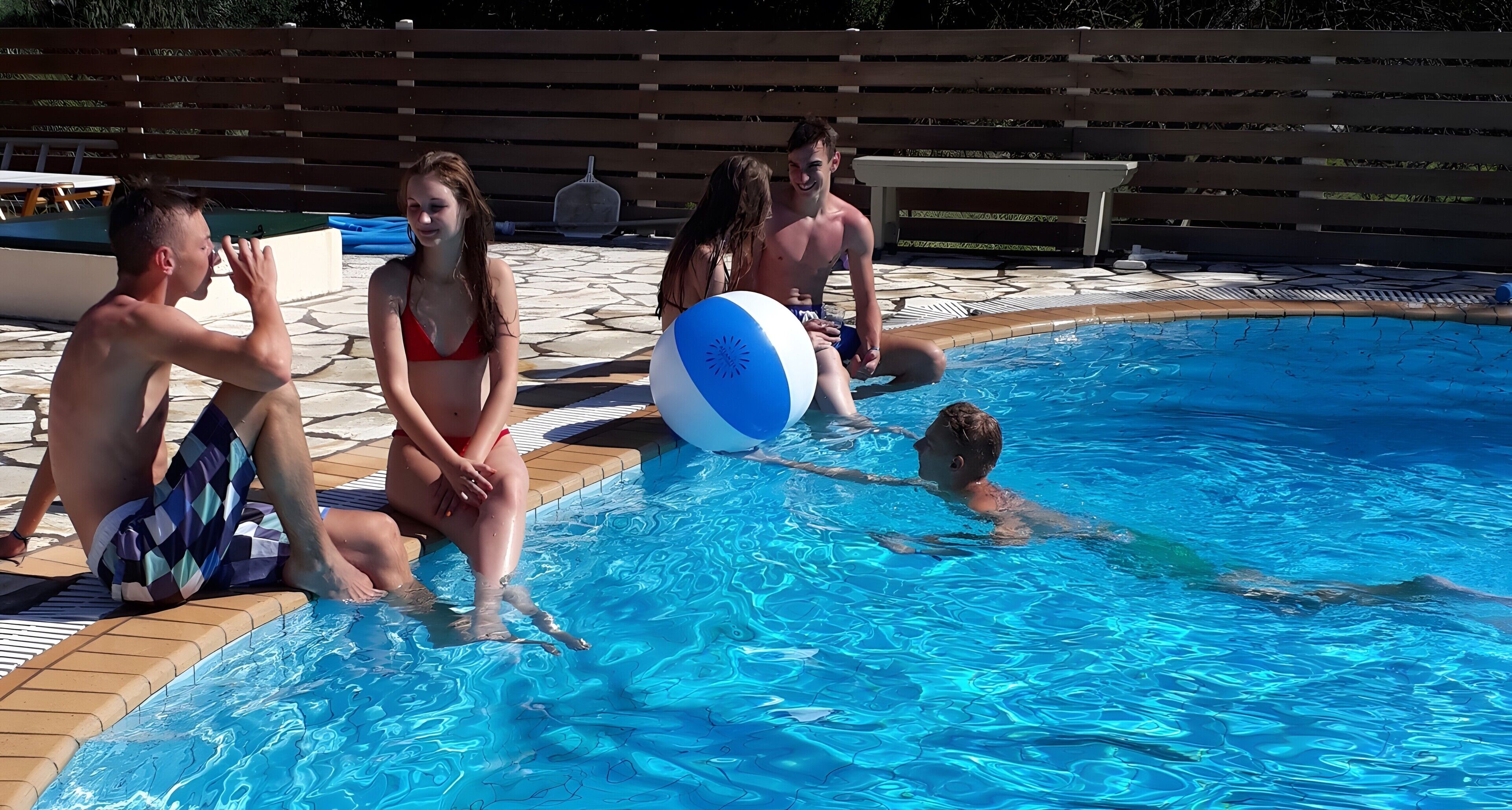 A swimming pool with people around it, a wooden deck, and a fence in the background.