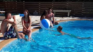 A swimming pool with people around it, a wooden deck, and a fence in the background.