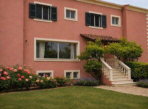 A pink building with multiple windows, a staircase, and a well-maintained garden with pink and green plants. - Poolfront Cabana 13 – Small Double Room by the Pool (Kontokali)