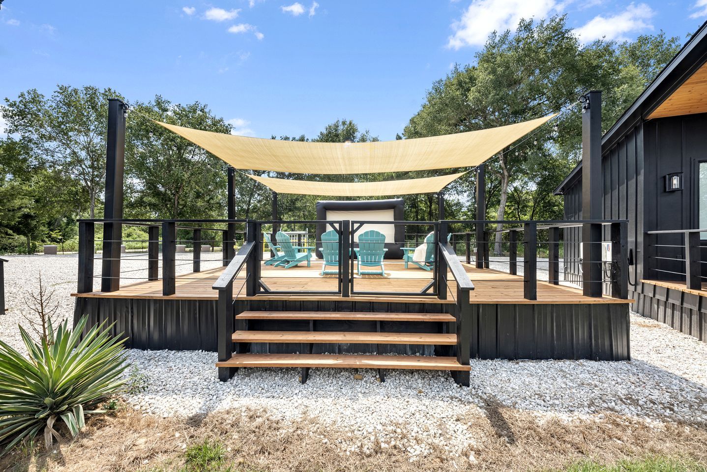 A wooden deck with steps leading to a seating area with chairs, a shaded canopy, and a building in the background.