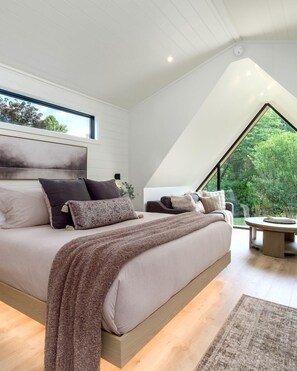 Ein modernes Schlafzimmer mit einem großen Bett, Blick ins Grüne durch ein Dachfenster und einem runden Tisch mit einem Stuhl.