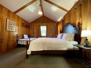 Un dormitorio con paneles de madera, una cama grande, una cama más pequeña, un escritorio con lámpara y una lámpara colgante.