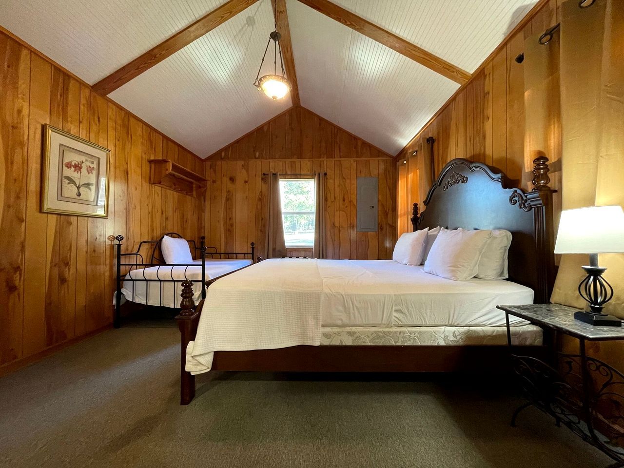 A wooden-paneled bedroom with a large bed, a smaller bed, a desk with a lamp, and a hanging light fixture.