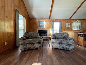 L’intérieur d’une cabane avec des murs en bois, une cheminée, deux canapés à motifs, une table à manger et un coin cuisine.