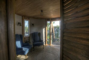 Une pièce aux murs en bois, avec deux fauteuils bleus, un petit tapis, une fenêtre et une porte donnant sur l’extérieur.