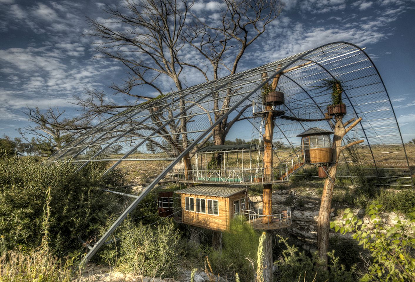 Une cabane dans les arbres reliée à un pont métallique, entourée d’arbres et de végétation.