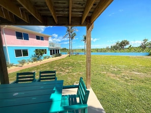 A covered outdoor seating area with a table and chairs, a building with a pink and blue facade, and a view of a body of water and palm trees in the background. - Fun Oceanfront 2 Bedroom Villa with Private Beach Access in Cocoa Beach (Cocoa Beach)