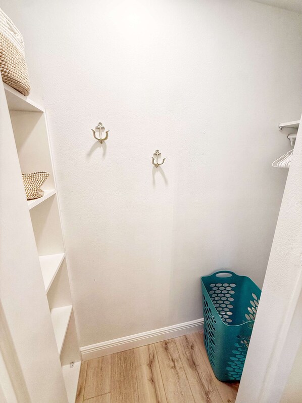 Una habitación pequeña con paredes blancas, dos ganchos en la pared, suelo de madera y una cesta color turquesa.