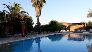 Une piscine dont les bords sont recouverts de carreaux, avec des chaises longues, des palmiers et un espace abrité comprenant des tables et des chaises.