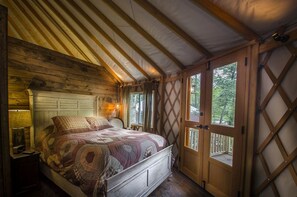 A cozy bedroom with a wooden ceiling, a bed with a patterned bedspread, a nightstand with a lamp, and a window with a lattice design.