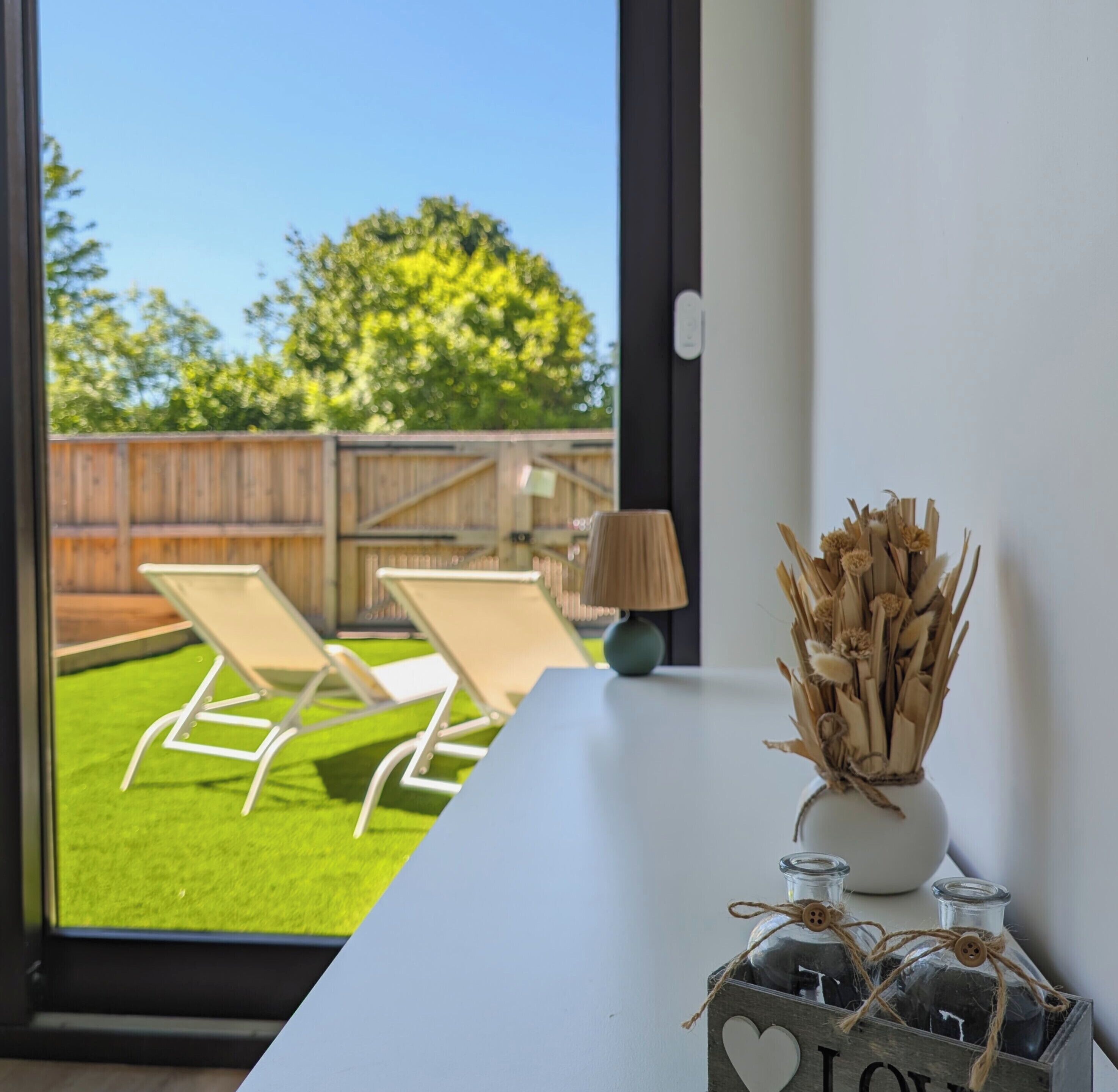 A room with a view of a backyard, a white table, a lamp, and a decorative vase with dried plants.