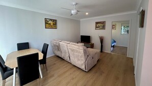 A living room with a sofa, a TV, a ceiling fan, and two paintings on the wall. - One Bedroom Blue Horizon close to everything (Tuncurry)
