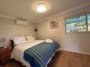 A bedroom with a bed, a nightstand, a lamp, a window with a view, and an air conditioning unit.