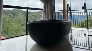 Un baño moderno con un lavabo negro grande, ventanas amplias con cortinas y vistas a un paisaje boscoso.