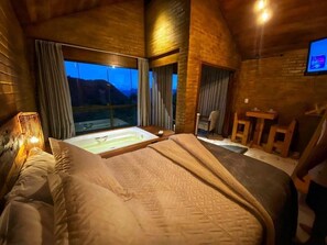 A bedroom with a large bed, a window with curtains, a small desk, and a TV mounted on the wall. - Vila Italiana Chalés (Santa Teresa)
